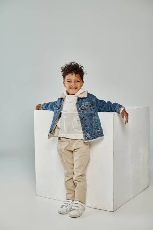 cheerful african american boy in winter attire and beanie sitting on concrete cube on grey backdrop