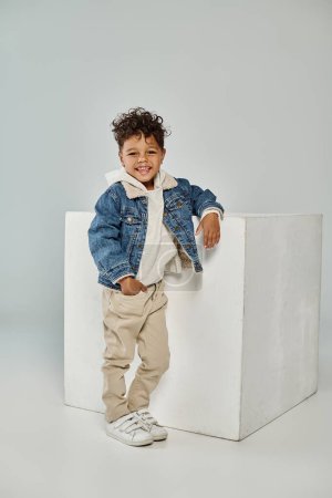 heureux garçon afro-américain en tenue d'hiver et bonnet assis sur cube de béton sur fond gris
