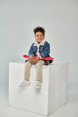 cheerful african american boy in winter attire sitting on cube with penny board on grey backdrop