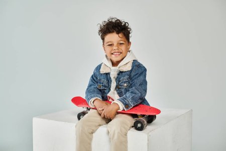 glücklicher afrikanisch-amerikanischer Junge in Winterkleidung sitzt auf Würfel mit Penny-Board auf grauem Hintergrund