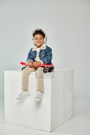 Téléchargez les photos : Mignon afro-américain garçon en tenue d'hiver et écouteurs assis sur cube avec panneau penny sur gris - en image libre de droit