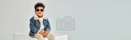 curly african american boy in winter attire and sunglasses sitting on cube on grey backdrop, banner