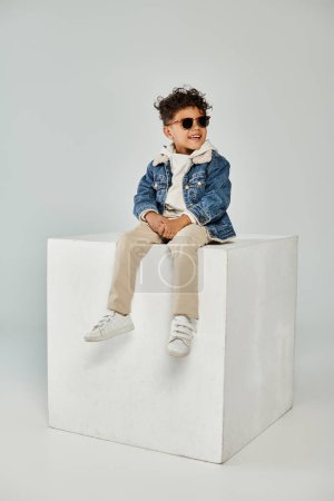 joyful curly african american boy in winter attire and sunglasses sitting on cube on grey backdrop