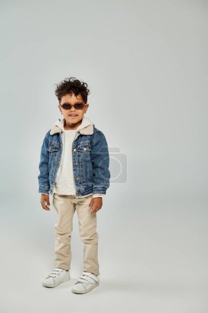 alegre rizado africano americano chico en invierno traje y gafas de sol de pie en gris telón de fondo
