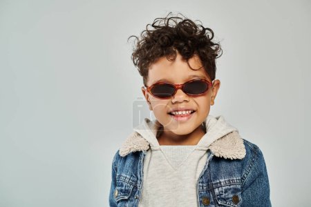 portrait, cheerful and curly african american boy in winter attire and sunglasses on grey backdrop