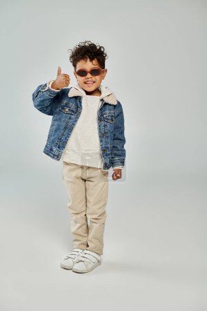 happy curly african american boy in winter attire and sunglasses showing thumb up on grey backdrop