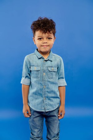 Téléchargez les photos : Portrait de heureux garçon afro-américain en costume de denim regardant la caméra sur fond bleu - en image libre de droit