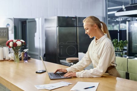 smiling blonde woman working on laptop near menu cards on counter in modern cafe, small business