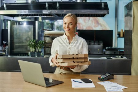Lächelnde blonde Frau mit Pizzakartons in der Nähe digitaler Geräte und Zahlungsterminal am Tresen im Café