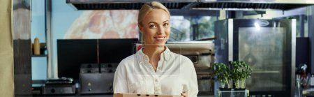 gérant blond joyeux tenant boîte à pizza et souriant à la caméra dans un café moderne, bannière horizontale