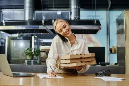 mujer rubia feliz con cajas de pizza hablando en el teléfono inteligente y el orden de escritura en el mostrador en la cafetería