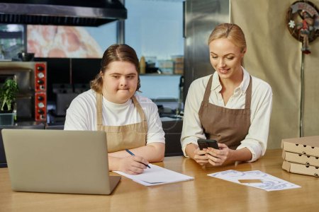 Junge Frau mit geistiger Behinderung schreibt Bestellung in der Nähe von Laptop und Manager mit Smartphone im Café