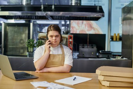 Téléchargez les photos : Jeune femme avec le syndrome du duvet travaillant dans un café et parlant sur smartphone près d'un ordinateur portable et des boîtes à pizza - en image libre de droit