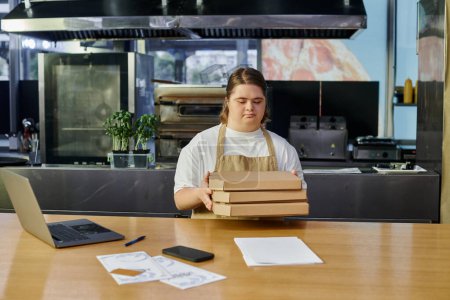 female employee with down syndrome holding pizza boxes near laptop and smartphone on counter in cafe