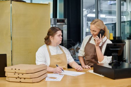 fröhliche Managerin spricht in modernem Café mit Smartphone neben Mitarbeiterin mit Down-Syndrom