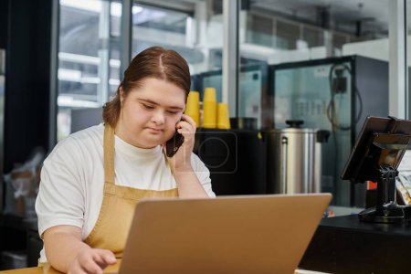 Mitarbeiterin mit Down-Syndrom spricht während der Arbeit im modernen Café auf dem Smartphone neben dem Laptop