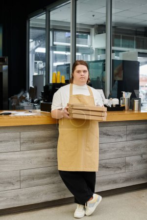Foto de Joven empleada con síndrome de Down de pie con cajas de pizza en el mostrador en la cafetería moderna - Imagen libre de derechos