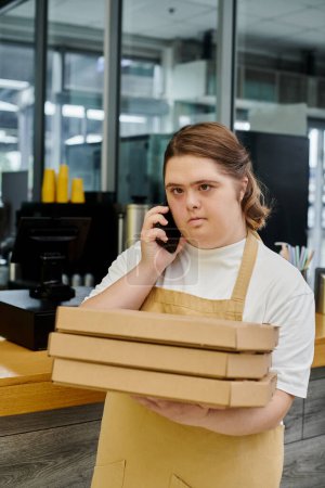 Téléchargez les photos : Jeune femme avec le syndrome du duvet tenant des boîtes à pizza et parlant sur smartphone tout en travaillant dans un café - en image libre de droit