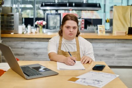 Junge Frau mit geistiger Behinderung schreibt Bestellung in der Nähe von Laptop und Smartphone auf Tisch im Café