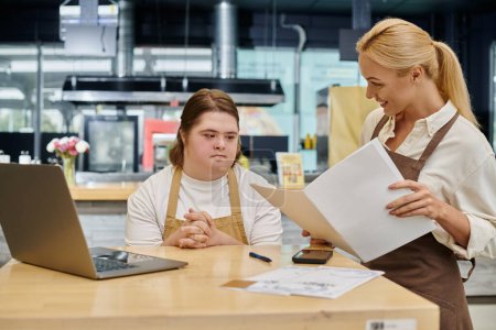 joyful administrator sitting with order book near woman with down syndrome at laptop in cafe