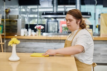 mujer joven con síndrome de Down limpiando mesa con trapo mientras trabaja en la cafetería moderna, inclusividad