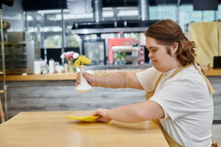 young woman with down syndrome holding vase with flowers and wiping table with rag in modern cafe