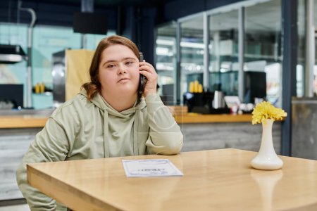 young woman with down syndrome talking on mobile phone on table in modern cafe, inclusivity
