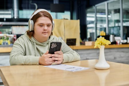 Frau mit Down-Syndrom hält Smartphone in der Hand und hört Musik über Kopfhörer in gemütlichem Café
