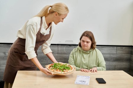 camarera sonriente propone deliciosa pizza a cliente femenino con síndrome de Down sentado en acogedora cafetería
