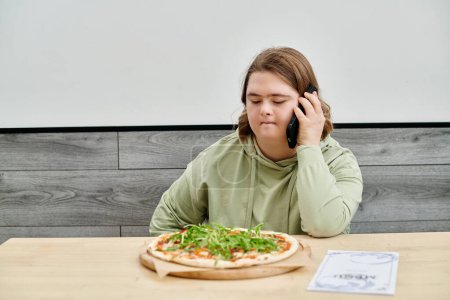 Junge Frau mit geistiger Störung spricht in modernem Café neben leckerer Pizza auf Smartphone