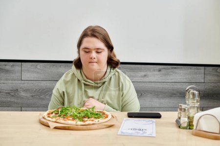 Foto de Mujer joven con síndrome de Down mirando deliciosa pizza mientras está sentado en la cafetería acogedora moderna - Imagen libre de derechos