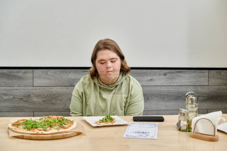 mujer joven con discapacidad mental mirando deliciosa pizza mientras está sentado en la cafetería acogedora moderna