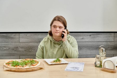 Junge Frau mit geistiger Störung spricht in modernem Café neben leckerer Pizza auf Smartphone