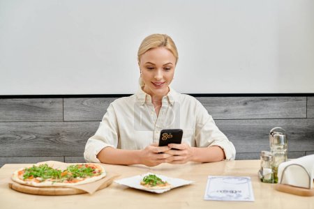 sonriente mujer rubia mensajería en el teléfono inteligente cerca de deliciosa pizza en la mesa en la cafetería acogedora moderna