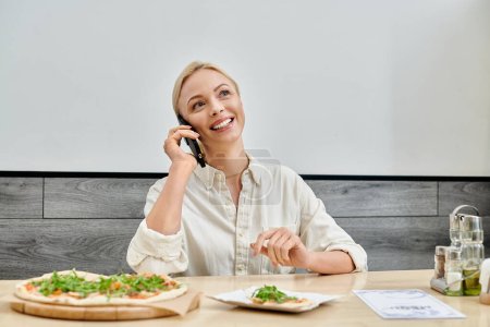 glücklich blonde Frau Nachrichten auf dem Smartphone in der Nähe leckere Pizza auf dem Tisch in modernen gemütlichen Café