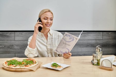 Lächelnde blonde Frau, die auf dem Smartphone spricht und in der Nähe einer leckeren Pizza im Café in die Speisekarte schaut