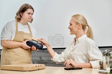 young waitress with mental disability holding payment terminal near pleased woman in modern cafe