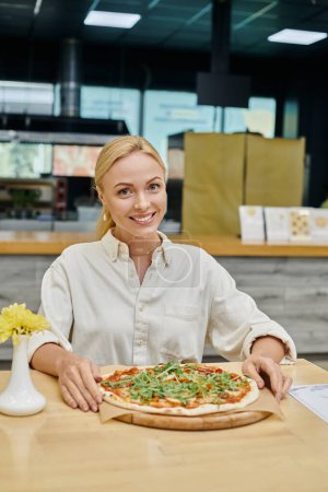 mujer rubia complacida sentada en la mesa cerca de deliciosa pizza en la cafetería moderna, delicia culinaria