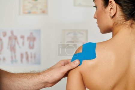 Téléchargez les photos : Vue arrière de la patiente attrayante avec la main du médecin qui met des bandes de kinesio sur son corps - en image libre de droit