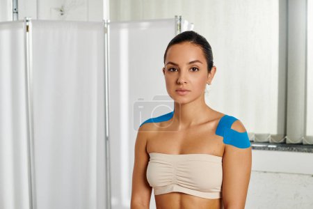 attraktive junge Patientin mit kinesiologischen Tapes auf den Schultern, die in die Kamera schaut, Gesundheitswesen