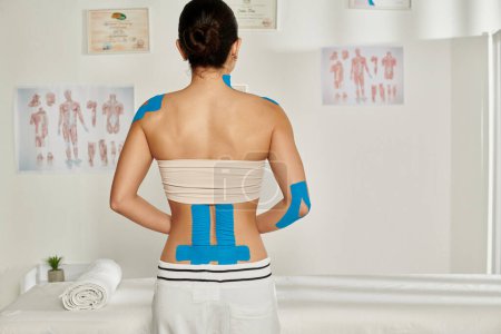 Rückenansicht einer jungen Patientin mit Kinesiobändern am Rücken und Ellbogen während des Termins