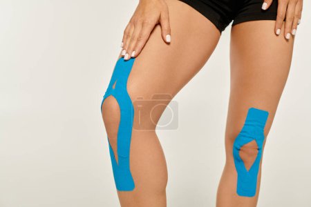 Foto de Vista recortada de mujer joven en ropa deportiva con cintas kinesiológicas en sus rodillas sobre fondo gris - Imagen libre de derechos