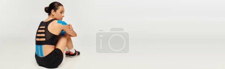 Téléchargez les photos : Belle jeune femme en tenue de sport assise sur le sol avec des bandes kinésiologiques sur le dos, bannière - en image libre de droit