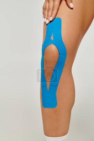 Foto de Vista recortada de mujer joven con cintas cinesiológicas azules en su rodilla posando sobre fondo gris - Imagen libre de derechos