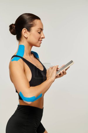 Foto de Hermosa mujer alegre en ropa deportiva negro mirando su teléfono con cintas kinesiológicas en su cuerpo - Imagen libre de derechos