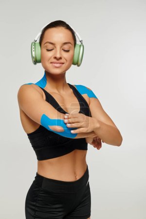 Foto de Atractiva mujer alegre en ropa deportiva con auriculares y cintas kinesiológicas en el codo y los hombros - Imagen libre de derechos