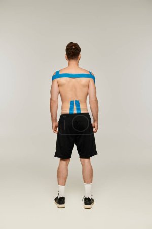 Foto de Vista trasera del hombre en pantalones deportivos negros con cintas kinesiológicas en su cuerpo sobre fondo gris - Imagen libre de derechos