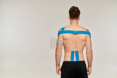 Foto de Vista posterior del hombre en pantalones deportivos con cintas kinesiológicas en la espalda y el hombro sobre fondo gris - Imagen libre de derechos