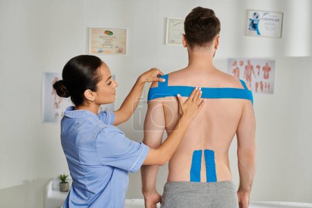 Photo for Beautiful young doctor in medical costume putting kinesio tapes on shoulders of her bearded patient - Royalty Free Image