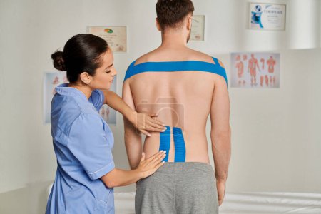Photo for Attractive young doctor in medical costume putting kinesiological tapes on back of her patient - Royalty Free Image